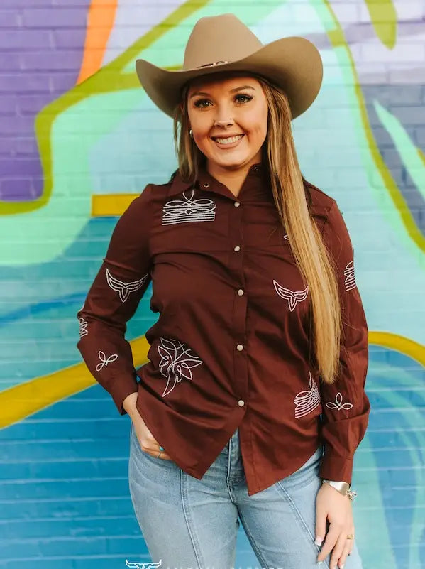 Woman in a deep brown pearl snap shirt with white western embroidery and a tan cowboy hat
